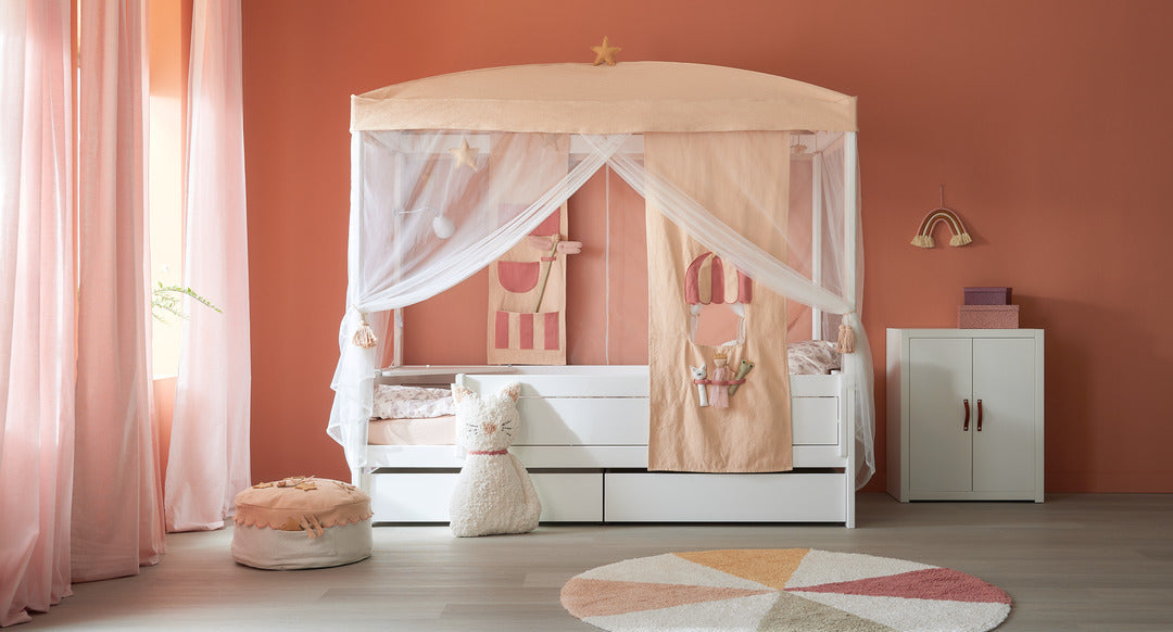 LIFETIME Kidsrooms children’s room with white canopy bed, fabric panels, plush toys, and storage cabinet, styled with soft pink curtains and a round pastel rug on wooden floor